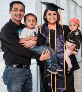 Female student in graduation regalia holding a baby. To the left, her husband holding a toddler